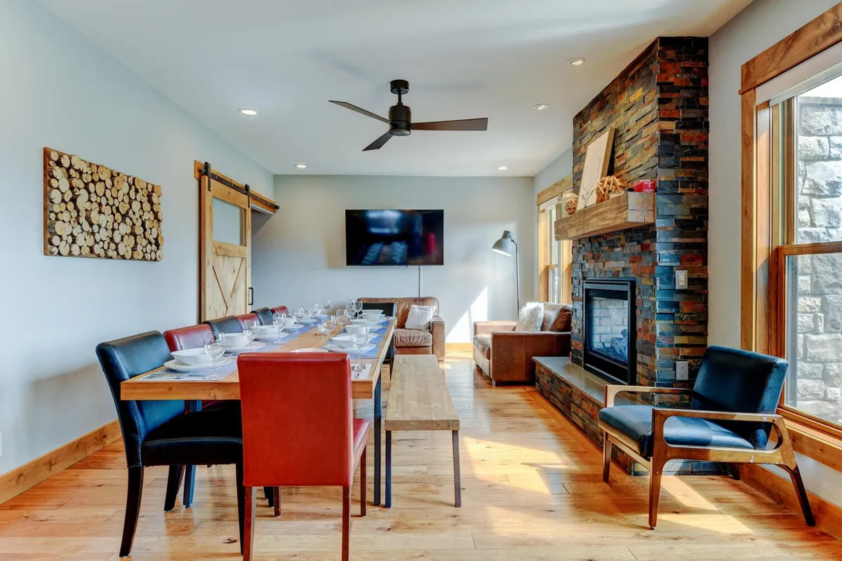 A warmly lit dining area with a long table, a cozy fireplace, a sliding barn door, and a wall-mounted television. The scene evokes family dinners, celebrations, and relaxed evenings watching TV by the fire.