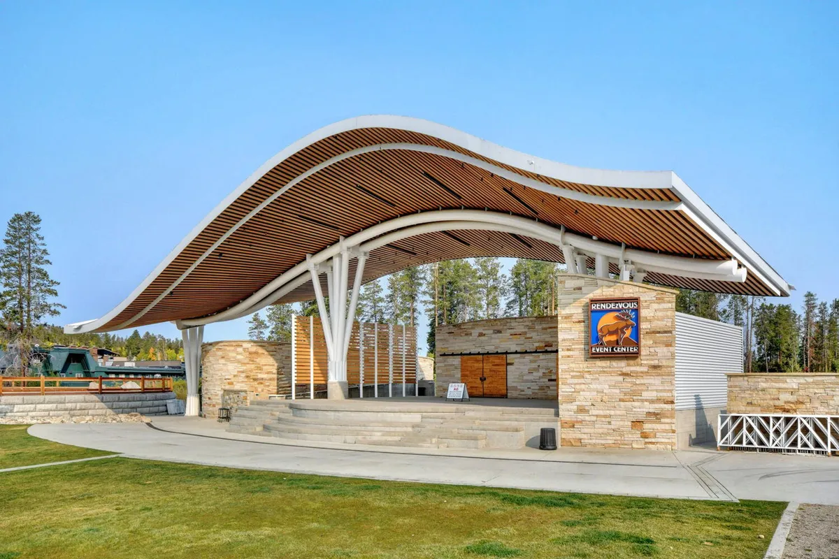 An outdoor event space is shown with a distinctive curved roof structure, stone pillars, wooden accents, and an open stage. The area is set for concerts or festivals, with open green space inviting gatherings and seasonal celebrations.