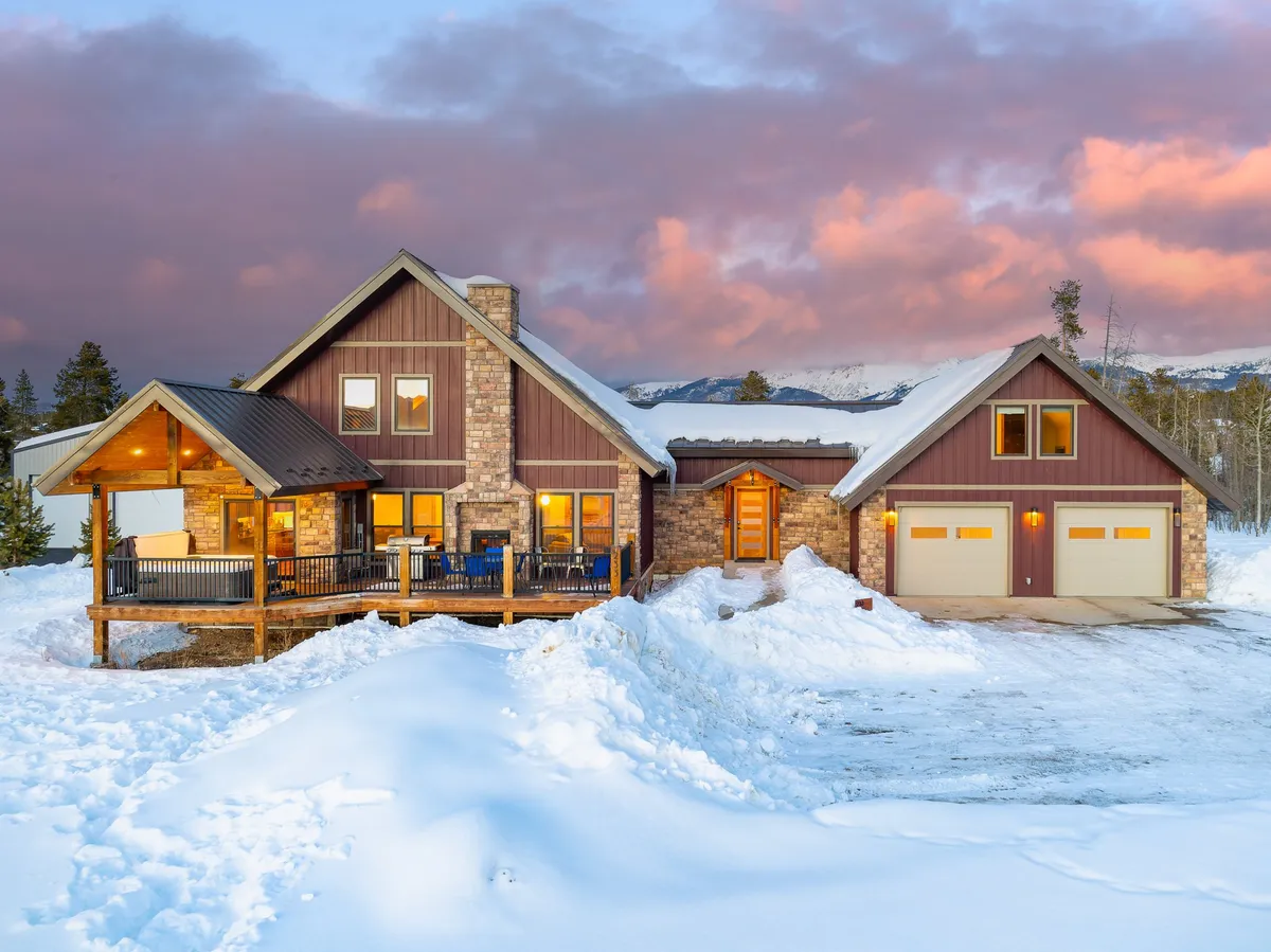 The image features the impressive stone and wood facade of the mountain home, with a welcoming porch set against a snow-covered landscape and sunset-lit clouds. The scene evokes a sense of warmth, adventure, and the unique blend of resort luxury and rugged outdoor beauty.