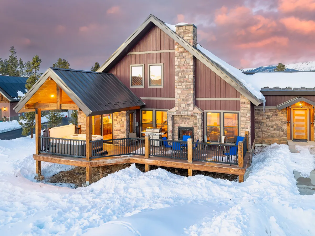 A wider view of the wooden deck featuring the hot tub, stone facade of the home, outdoor seating, and large windows. The setting transitions from day to night, inviting stargazing or cozy moments with a warm beverage, and highlights how the outdoor living space seamlessly connects with the interior.