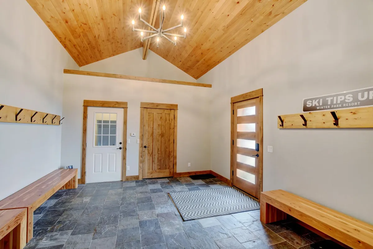This image showcases a warm and functional mudroom featuring cozy wooden benches, slate flooring, a modern light fixture, a wooden door with glass panels, and wall hooks specifically designed for ski gear. The space is clearly equipped for guests returning from the slopes, offering seating for removing boots and organized storage for outerwear.