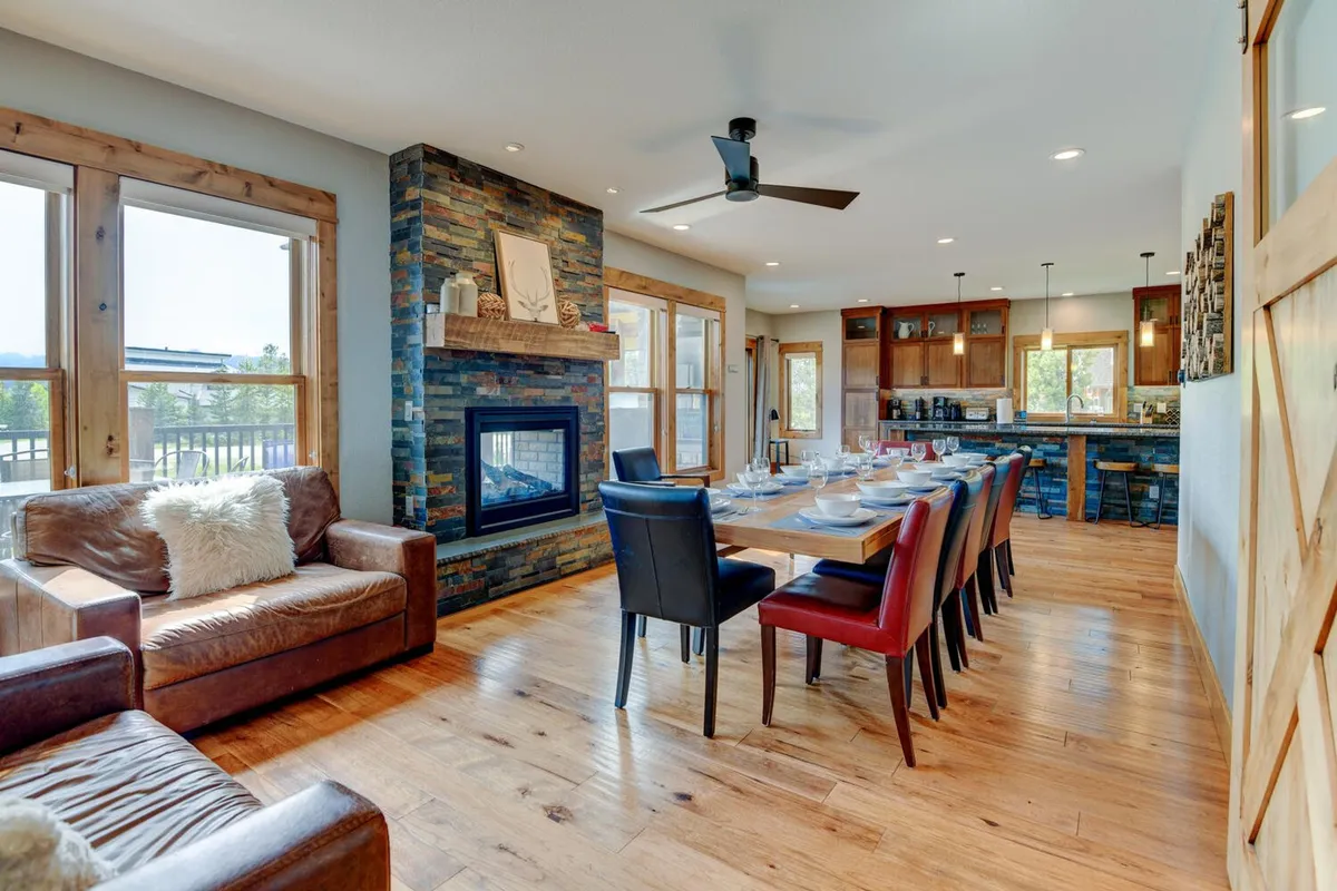 A warm, inviting great room with a stone gas fireplace, a large dining table set for a meal, expansive windows framing views of Byers Peak, and exposed wooden beams overhead. The space exudes comfort, with sunlight streaming in and abundant seating for gatherings.