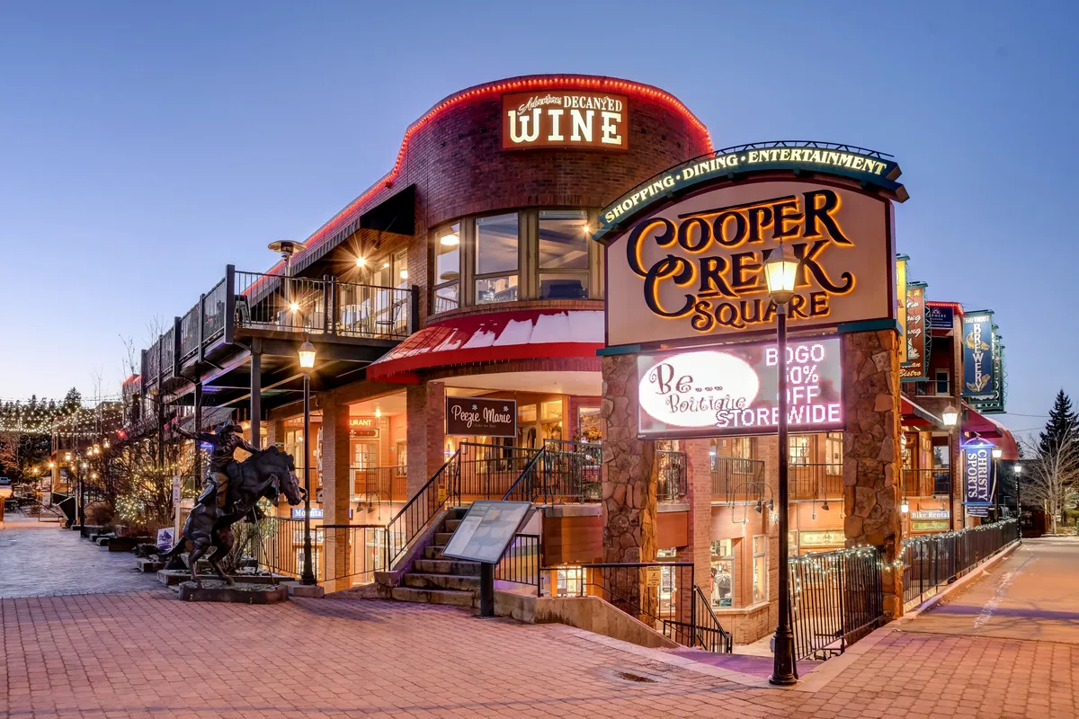 The vibrant exterior of Cooper Creek Square is shown with a prominent sign, a wine shop, decorative lighting, and an inviting outdoor seating area. The lively scene hints at the nearby dining, shopping, and social experiences available in downtown Winter Park.