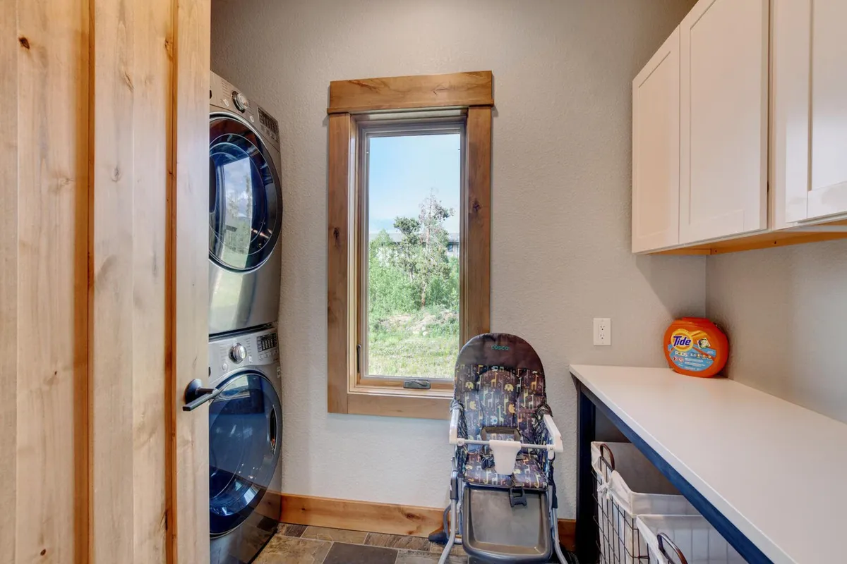 A practical laundry area featuring a stacked washer and dryer next to a sunlit window, alongside a child's high chair and organized laundry supplies. This scene blends utility and family-friendliness, underscoring thoughtful touches like a booster seat and easy access for families with young children.
