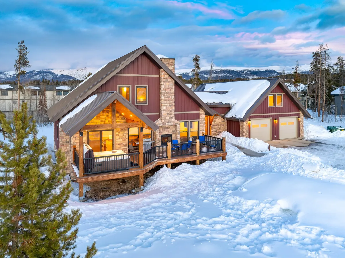 The hero image showcases a stunning mountain home with a snow-covered roof, an inviting wooden deck, and a striking stone facade. The deck overlooks panoramic views of snow-capped mountains, with ample space for family gatherings, enjoying hot cocoa, or watching the sunset. The yard below invites guests to build snowmen or gather around the fire pit, all against a backdrop of Colorado's breathtaking winter scenery.