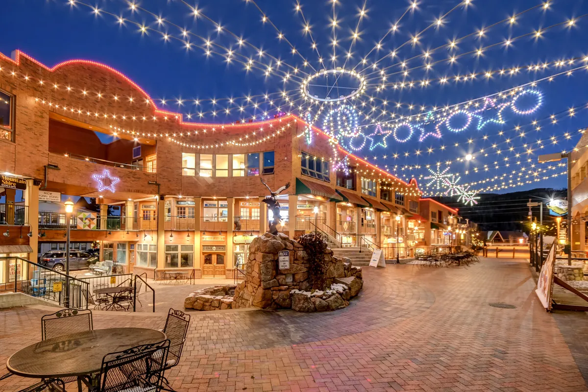 The image captures a lively downtown scene at dusk, featuring decorative storefronts illuminated by string lights and a central fountain sculpture. People are seen strolling, shopping, and dining outdoors, creating an atmosphere of year-round vibrancy and community spirit.